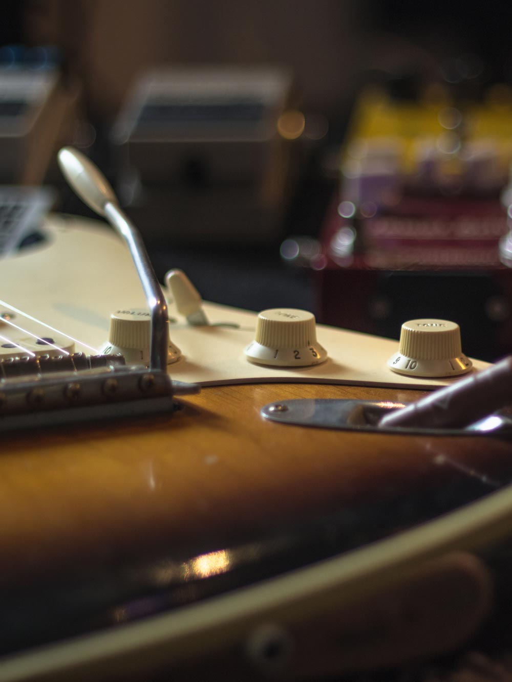 A close up shot of the switch, tone and volume controls on a Fender stratocaster. The image shows out of focus pedals in the back ground.